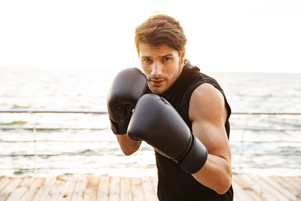 Foto de homem caucasiano em traje de treino trabalhando em luvas de boxe preto no cais de madeira à beira-mar — Fotografia de Stock