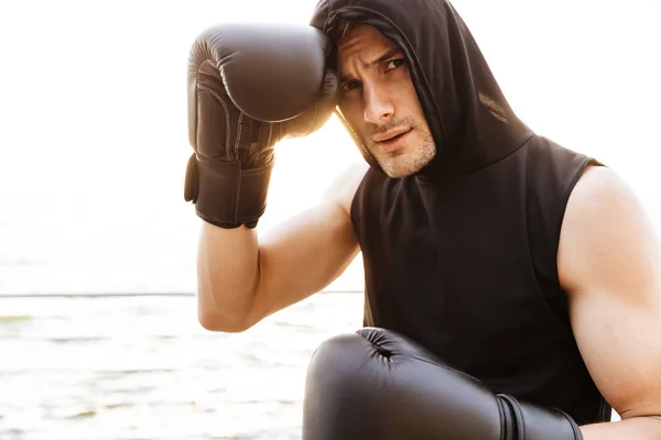 Foto de un joven con sudadera con capucha haciendo ejercicio con guantes de boxeo negros en un muelle de madera en la playa — Foto de Stock