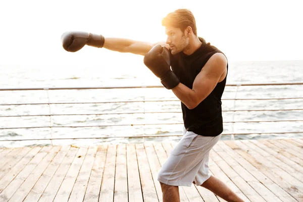 Foto de un joven en chándal haciendo ejercicio con guantes de boxeo negros en un muelle de madera en la playa — Foto de Stock