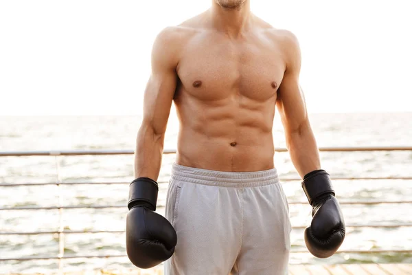 Foto recortada fuerte hombre semidesnudo haciendo ejercicio en guantes de boxeo negro en el muelle de madera en la playa —  Fotos de Stock