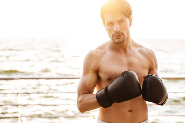 Foto de un hombre semidesnudo confiado mirando la cámara mientras hace ejercicio con guantes de boxeo negros en el muelle de madera en la playa —  Fotos de Stock