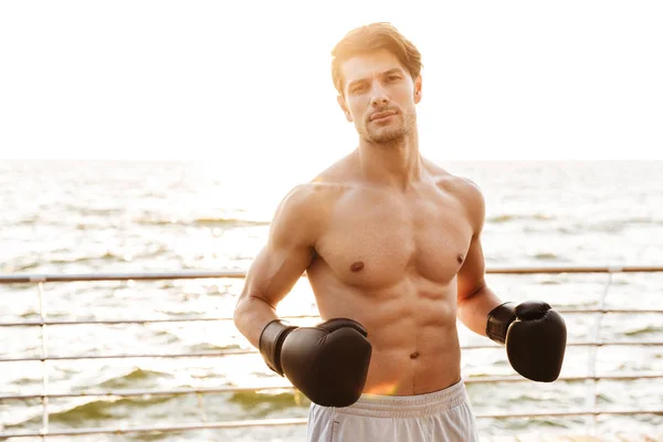 Foto de hombre semidesnudo satisfecho mirando la cámara mientras hace ejercicio con guantes de boxeo negros en el muelle de madera en la playa — Foto de Stock
