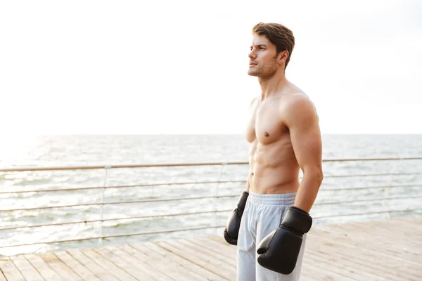 Foto de hombre semidesnudo satisfecho mirando a un lado mientras hace ejercicio en guantes de boxeo negro en el muelle de madera en la playa —  Fotos de Stock