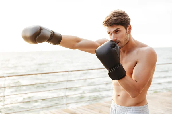 Photo of brunette half-naked man working out in black boxing gloves on wooden pier at seaside — Stock Photo, Image