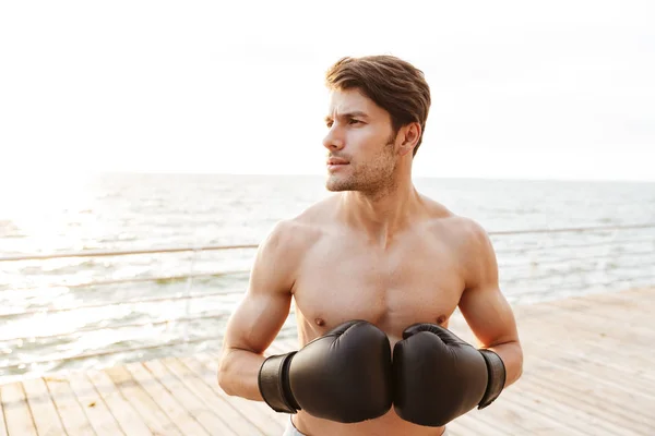 Foto eines zufriedenen halbnackten Mannes, der zur Seite schaut, während er in schwarzen Boxhandschuhen auf einem Holzsteg am Meeresufer trainiert — Stockfoto