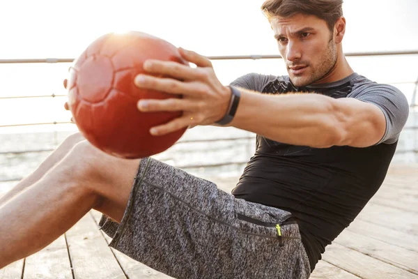 Foto de un hombre enfocado en chándal haciendo ejercicio con una pelota de fitness roja mientras hace ejercicio en un muelle de madera en la playa — Foto de Stock