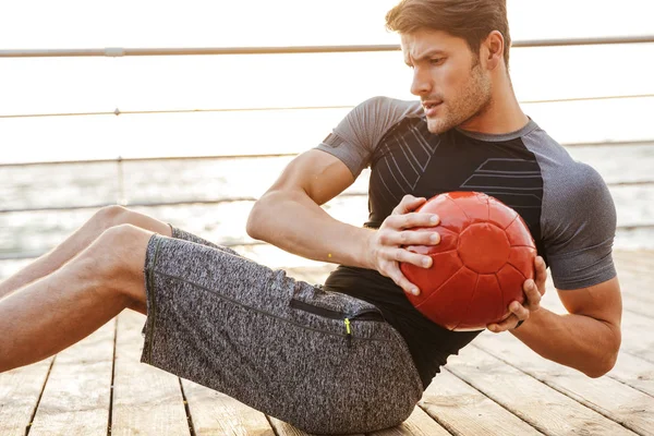 Foto av koncentrerad man i träningsoverall gör övning med röd Fitness boll medan du tränar på träbrygga vid havet — Stockfoto