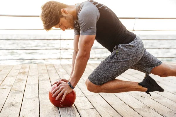 Foto de un hombre guapo en chándal haciendo ejercicio con una pelota de fitness roja mientras hace ejercicio en un muelle de madera en la playa — Foto de Stock