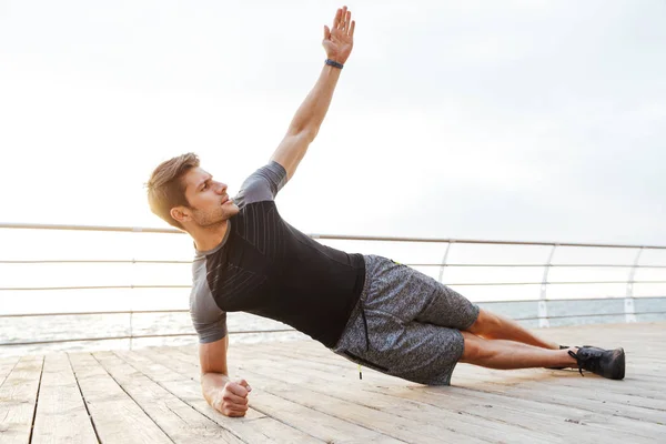 Foto de um homem forte em traje de treino levantando braço e fazendo prancha lateral enquanto trabalhava no cais de madeira à beira-mar — Fotografia de Stock