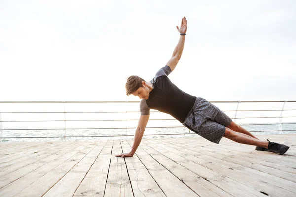 Foto de un hombre deportivo en chándal levantando el brazo y haciendo tablón lateral mientras hace ejercicio en el muelle de madera en la playa —  Fotos de Stock