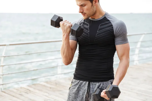 Serious concentrated young sports man outdoors at beach make exercises for upper arms with dumbbells. — Stock Photo, Image