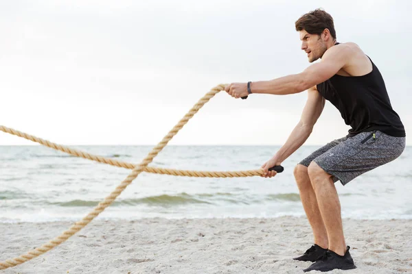 Φωτογραφία του ισχυρού νεαρό άντρα σε φόρμα εργασίας με σχοινί γυμναστικής στην παραλία στη θάλασσα — Φωτογραφία Αρχείου