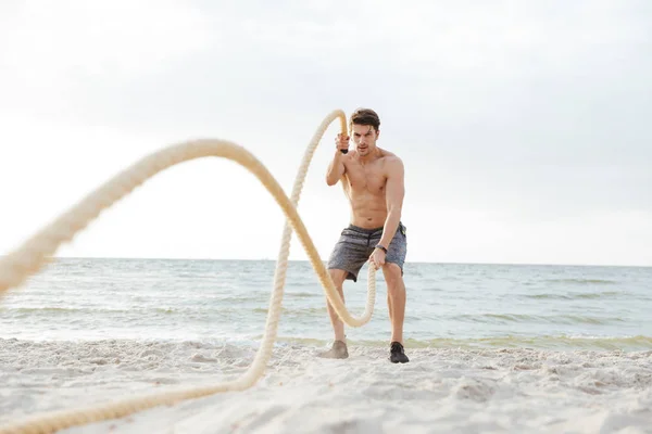 Fotografie silného polonahého muže, který pracuje s fitness lanem na pláži na pobřeží — Stock fotografie