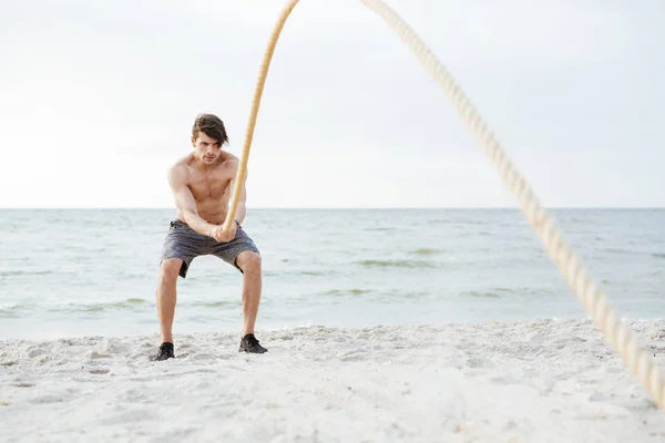 Fotografie brutálního napůl nahého muže, který pracuje s fitness lanem na pláži na pobřeží — Stock fotografie