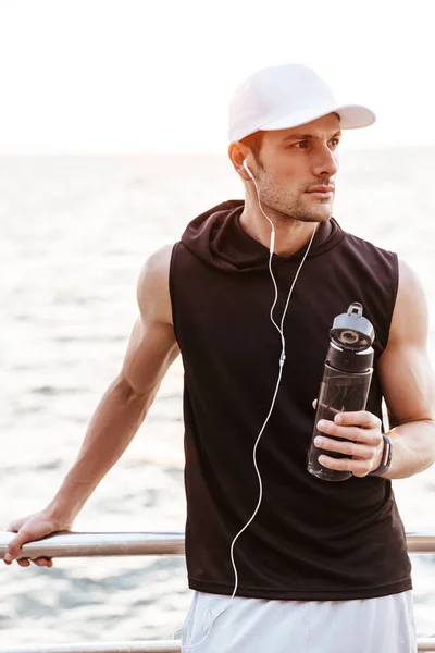 海辺の桟橋を歩きながらイヤホンと飲料水を使った白い帽子をかぶった真剣な男性の写真 — ストック写真