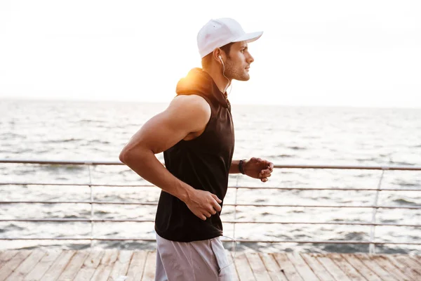 Foto de hombre caucásico en gorra blanca escuchando música con auriculares mientras trota en el muelle de madera en la playa — Foto de Stock