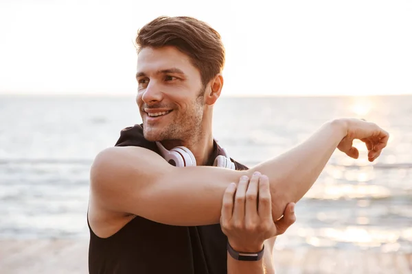 Photo smiling man in tracksuit with headphones doing exercise while working out at seaside — Stock Photo, Image