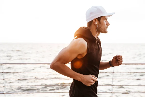 Foto de hombre atlético en gorra blanca escuchando música con auriculares mientras trota en el muelle de madera en la playa —  Fotos de Stock