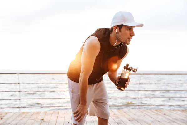 Φωτογραφία από σπορ άνθρωπος σε λευκό καπάκι ανάπαυσης και πόσιμου νερού, ενώ τρέξιμο σε ξύλινη προβλήτα στην παραλία — Φωτογραφία Αρχείου