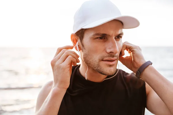Foto de hombre deportivo en gorra blanca escuchando música con auriculares mientras camina en la playa i — Foto de Stock
