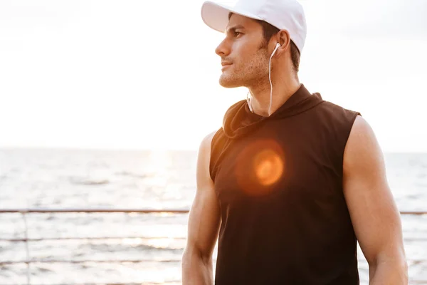 Foto de un hombre confiado en gorra blanca escuchando música con auriculares mientras camina a la orilla del mar — Foto de Stock