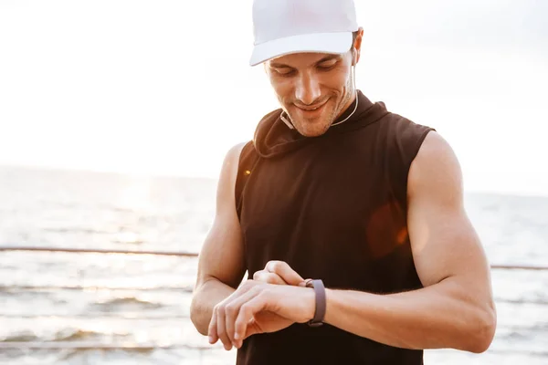 Foto de un hombre sonriente con gorra blanca escuchando música con auriculares y usando un reloj inteligente mientras camina a la orilla del mar — Foto de Stock