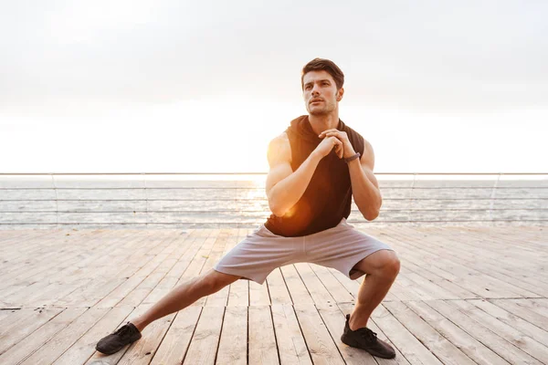 Foto de un joven en chándal haciendo ejercicio mientras hace ejercicio en el muelle de madera en la playa — Foto de Stock