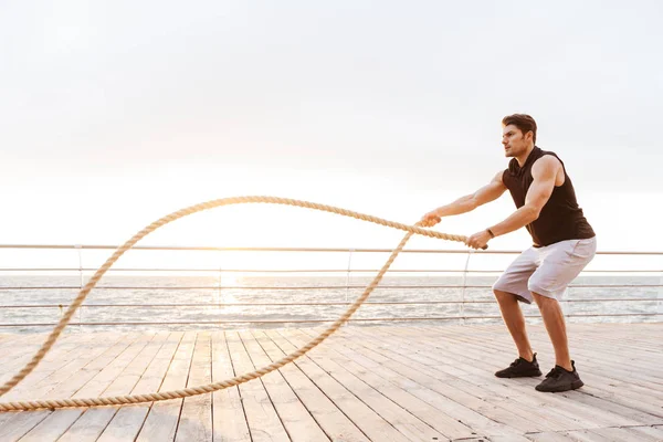 Fotó az erős ember melegítőben dolgoznak ki fitness kötél fából készült mólón a tengerparton — Stock Fotó