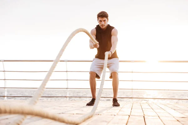 Deniz kenarında ahşap iskeleüzerinde fitness ipi ile çalışan eşofman odaklanmış adam Fotoğrafı — Stok fotoğraf