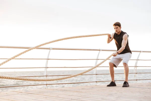 Foto de un hombre sin afeitar en chándal haciendo ejercicio con una cuerda de fitness en un muelle de madera en la playa — Foto de Stock