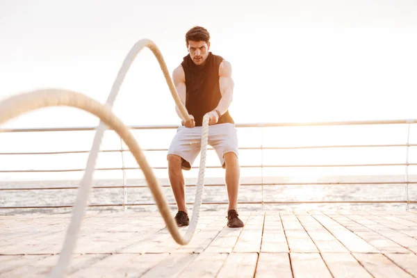 Foto de un hombre concentrado en chándal haciendo ejercicio con una cuerda de fitness en un muelle de madera en la playa —  Fotos de Stock