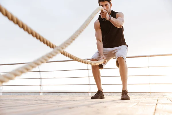 Foto de un hombre deportivo en chándal haciendo ejercicio con una cuerda de fitness en un muelle de madera en la playa — Foto de Stock
