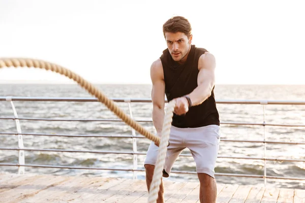 Foto de un hombre brutal en chándal haciendo ejercicio con una cuerda de fitness en un muelle de madera en la playa — Foto de Stock