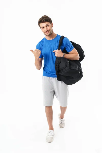 Blij optimistische knappe jonge sport fitness man staande geïsoleerd over witte muur achtergrond Holding tas wijzend naar copyspace. — Stockfoto
