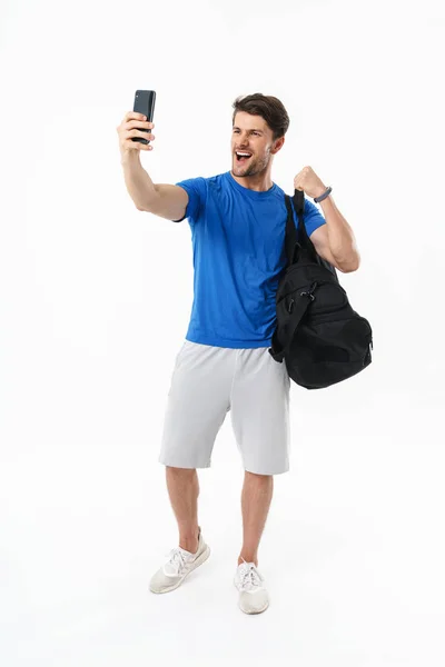 Émotionnel beau jeune sportif fitness homme debout isolé sur fond blanc sac de maintien faire selfie par téléphone mobile montrant biceps . — Photo