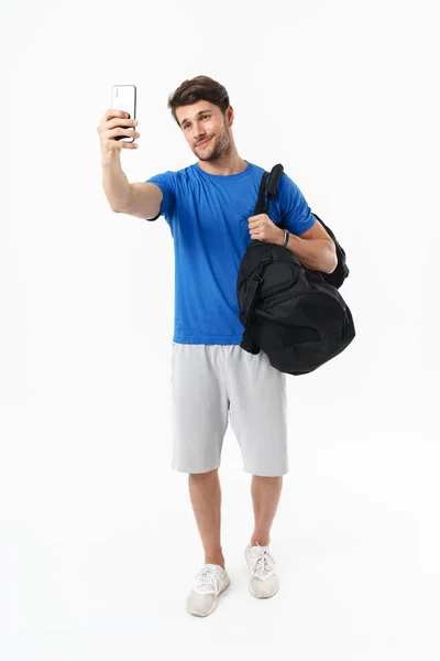 Zadowolony uśmiechnięty przystojny młody Sport fitness człowiek stojący na białym tle na biały ściana trzyma torba zrobić selfie przez telefon komórkowy. — Zdjęcie stockowe