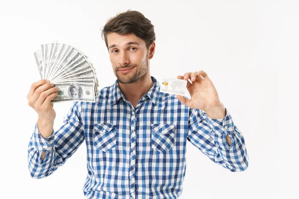 Bonito barbudo vestido com camisa casual posando isolado sobre fundo parede branca segurando dinheiro e cartão de crédito . — Fotografia de Stock