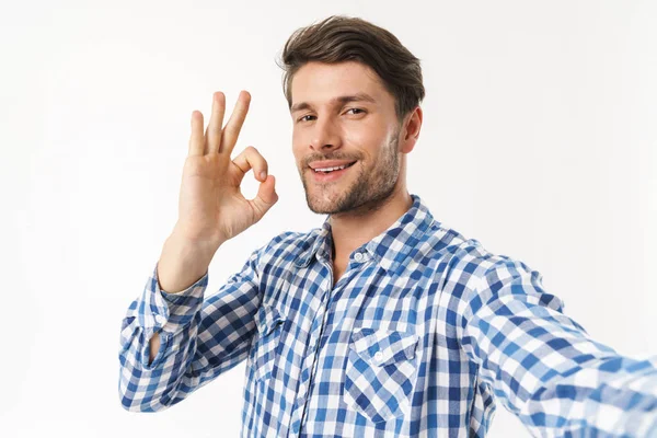 Happy handsome young man standing isolated over white wall background dressed in casual shirt make selfie by camera showing okay gesture. — Stock Photo, Image