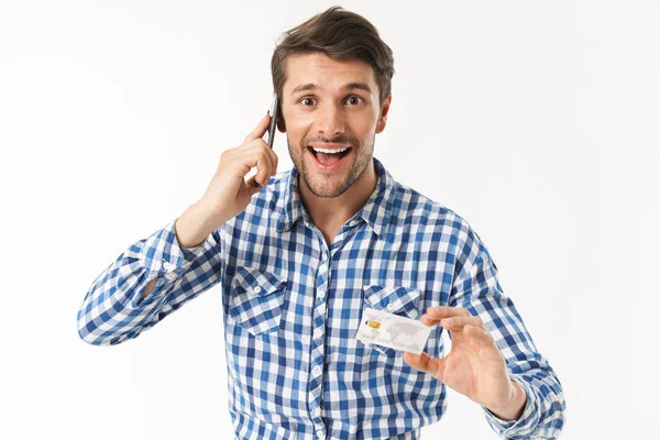 Emocionado joven guapo alegre de pie aislado sobre fondo blanco de la pared vestido con camisa casual hablando por teléfono móvil con tarjeta de crédito . — Foto de Stock