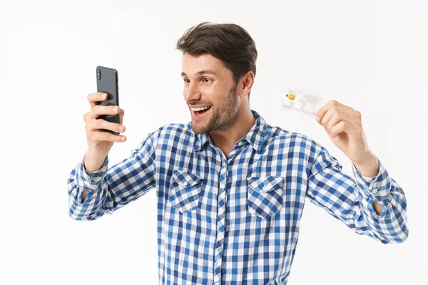 Meglepett csinos fiatalember álló izolált fehér fal háttér öltözött alkalmi inget használ mobiltelefon Holding hitelkártya. — Stock Fotó