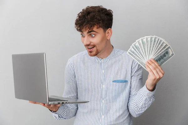 Портрет восторженного кавказца, одетого в рубашку, улыбающегося в камеру, держащего в руках серебряный ноутбук и кучу денег — стоковое фото