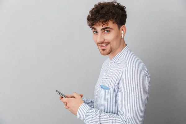 Imagen de un joven alegre con auriculares sonriendo a la cámara y sosteniendo el teléfono celular — Foto de Stock