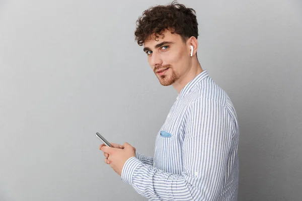 Imagen de un joven sin afeitar con auriculares mirando a la cámara y sosteniendo el teléfono celular — Foto de Stock