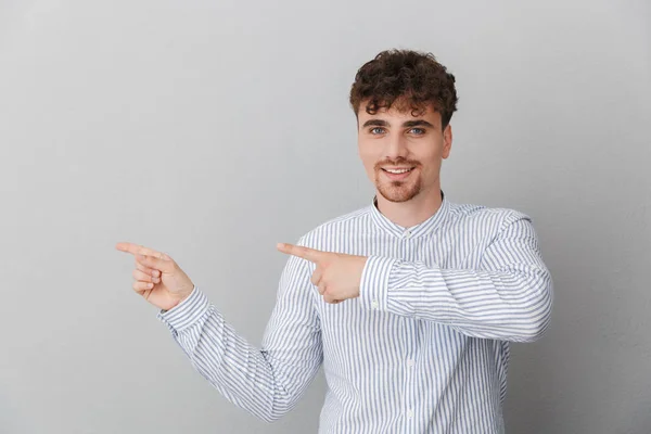 Portrait d'homme bouclé attrayant vêtu d'une chemise souriant à la caméra et pointant les doigts de côté à l'espace de copie — Photo