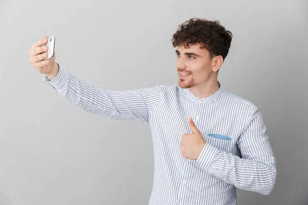 Retrato de un hombre hermoso y positivo vestido con camisa sonriendo mientras sostiene y toma una foto de selfie en un teléfono inteligente — Foto de Stock