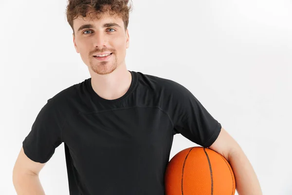 Фотография привлекательного молодого человека в футболке, улыбающегося и держащего мяч во время игры в баскетбол — стоковое фото