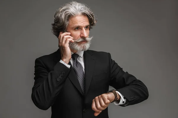 Bild eines selbstbewussten erwachsenen Geschäftsmannes im formalen schwarzen Anzug, der auf seinem Smartphone spricht und auf seine Armbanduhr blickt — Stockfoto