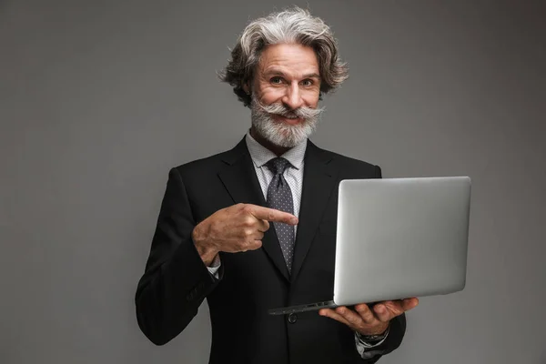 Imagen del alegre hombre de negocios adulto vestido con traje negro formal sonriendo y señalando con el dedo a la computadora portátil de plata —  Fotos de Stock