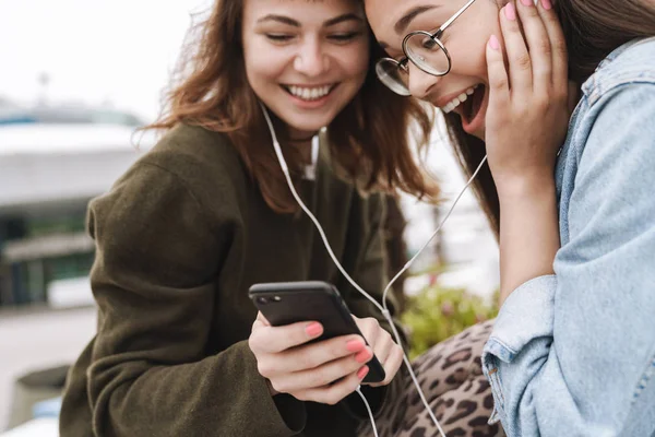 Megdöbbentette az érzelmi fiatal csinos barátok nők diákok séta szabadban hallgat zenét fülhallgató segítségével mobiltelefon. — Stock Fotó