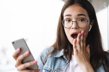 Cep telefonu sohbet kullanarak kulaklıkla müzik dinleyerek açık havada yürüyen gözlük takan heyecanlı genç güzel kadın öğrenci şok.